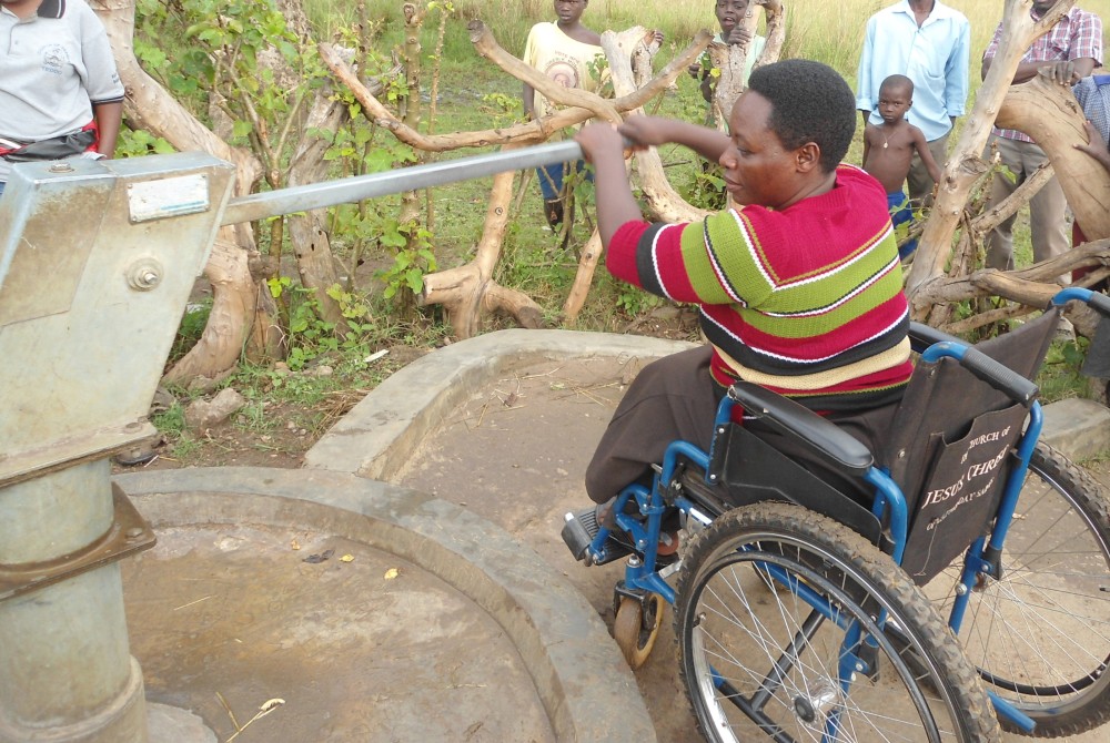 A woman using a wheelchair is operating a handpump from a concrete apron