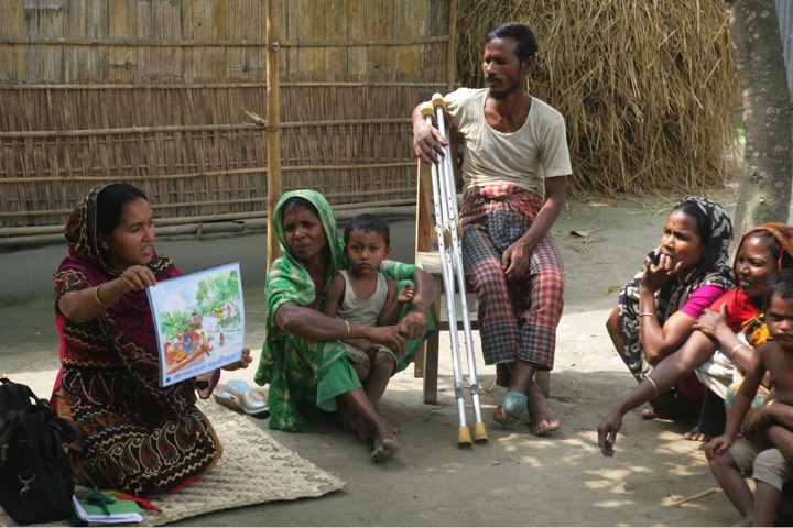 A woman sitting on the ground showing a poster of a mockdrill exercise for a group of women with children and a man sitting on a chair with two crutches resting on his legs