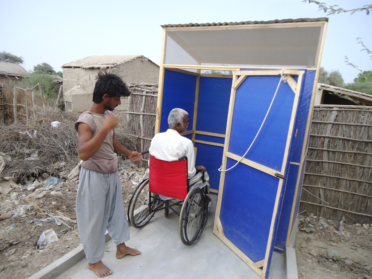 A man using a wheelchair is entering a latrine made from plastic tarps. The lartine is accessed by a ramp and the door has a lock function using a rope